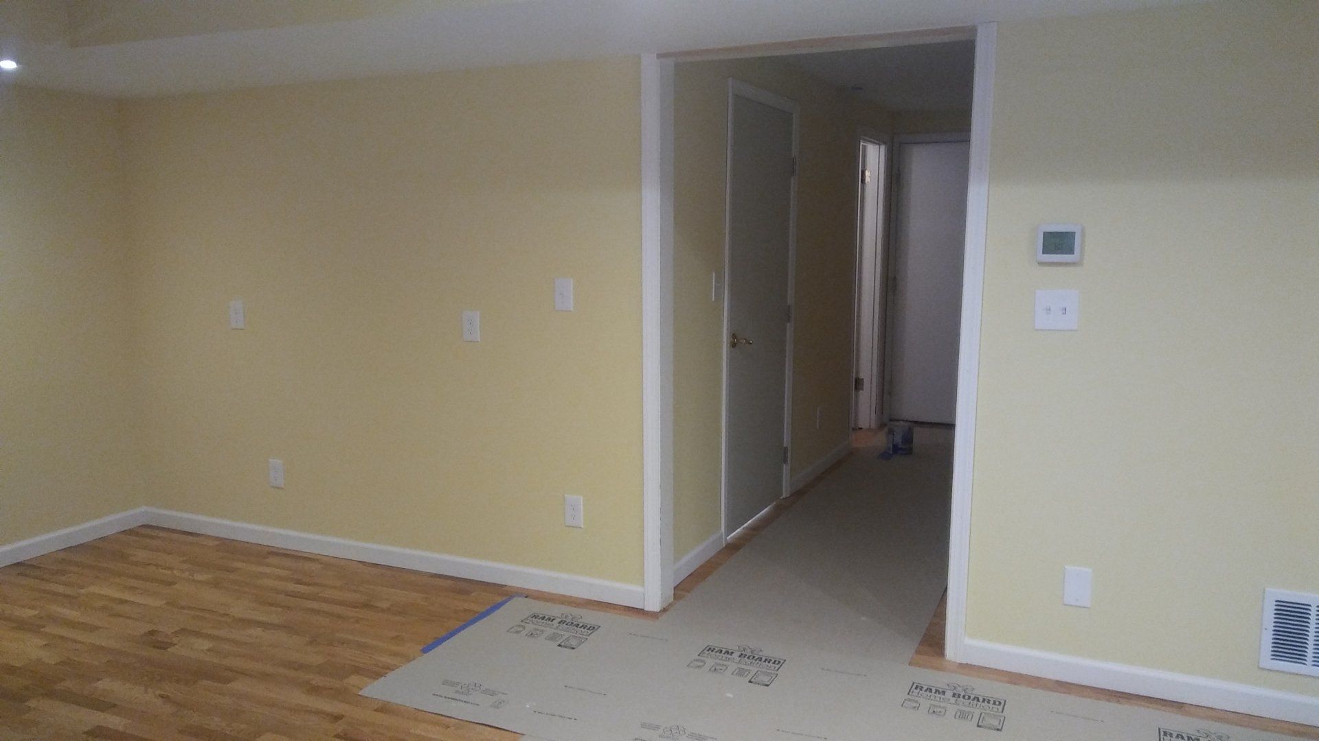 Empty beige living room with hardwood floor, hallway opening, and thermostat on the wall