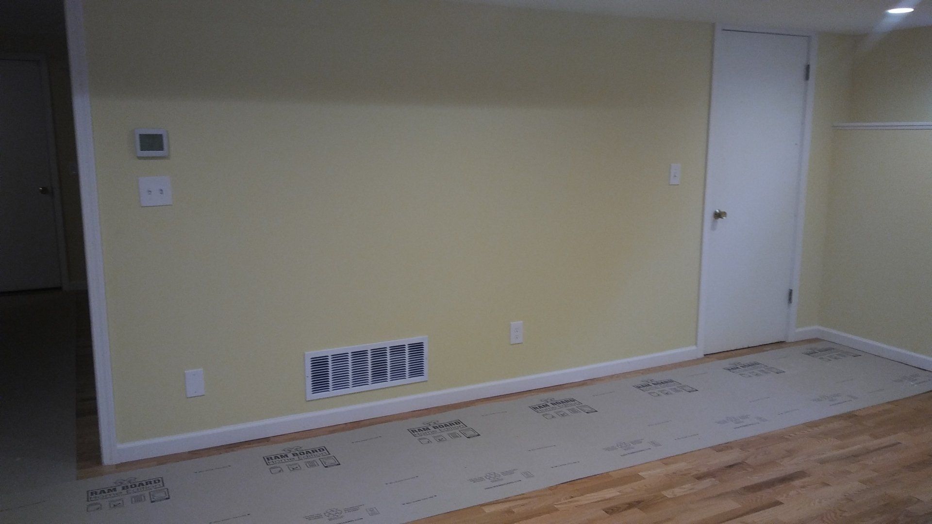 Empty beige room with baseboard vent, wall thermostat, and hardwood floor under renovation