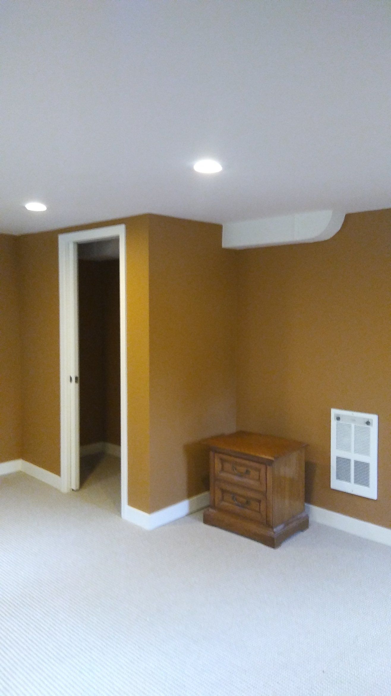 Small room with mustard walls, white carpet, recessed lights, doorway, and a wooden cabinet by a vent.