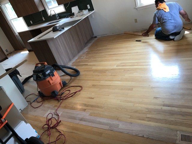 Person kneeling on hardwood floor beside vacuum, cleaning a kitchen area with cabinets and counter.