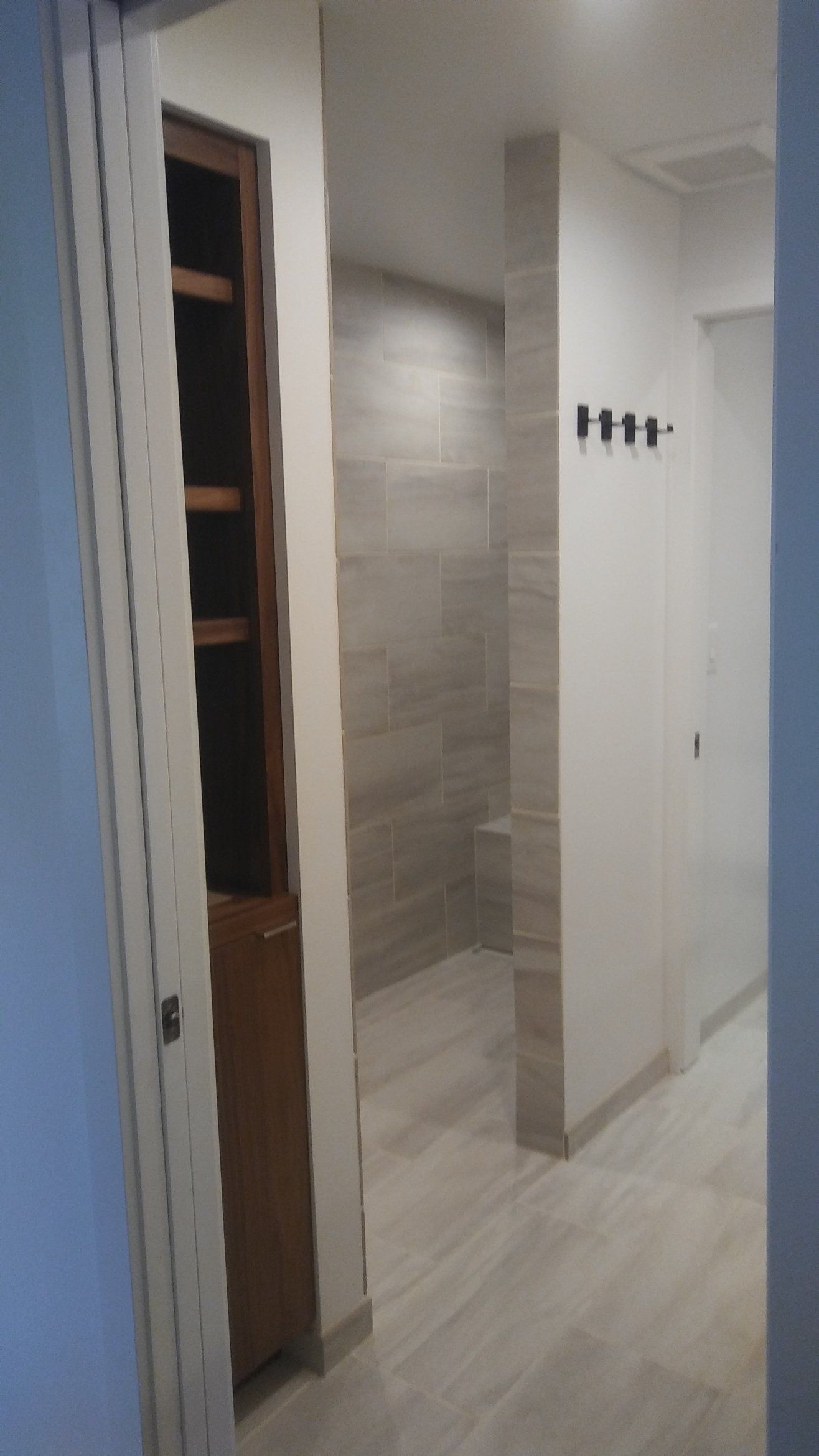 Narrow hallway with light tile flooring, white walls, and a doorway leading to a bathroom area.