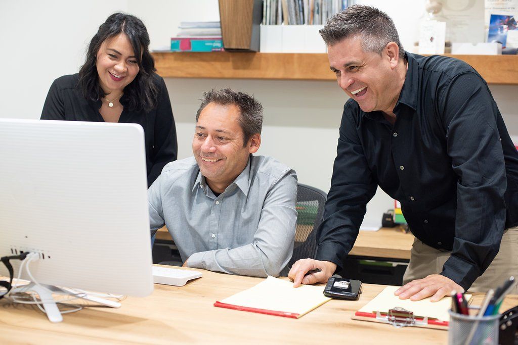 Three coworkers smiling around a desktop computer in an office meeting
