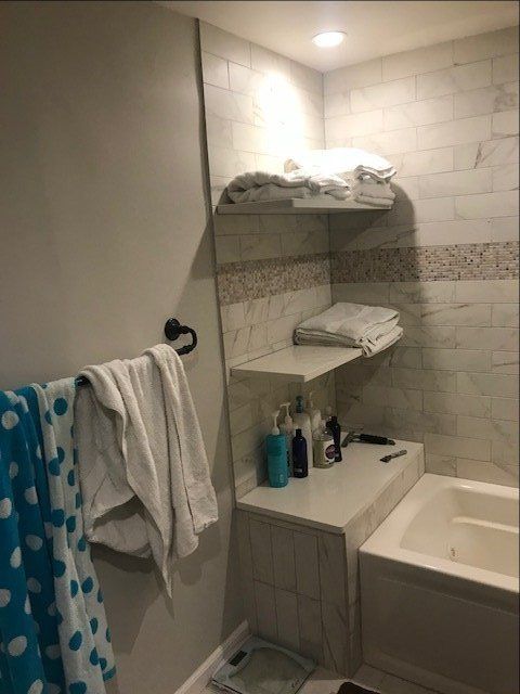 Small bathroom with shower-tub, shelves, towels, and toiletries in neutral tile.