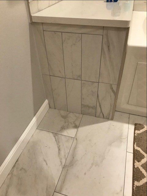 Bathroom corner with light gray tile wall, white marble-look floor, and small sink base cabinet