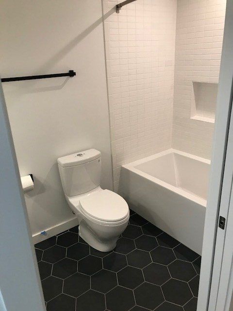 Small modern bathroom with a toilet, bathtub, white tile walls, and hexagonal dark floor tiles.