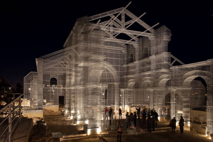 A wireframe sculpture of a building, illuminated at night, with a small group of people gathered.