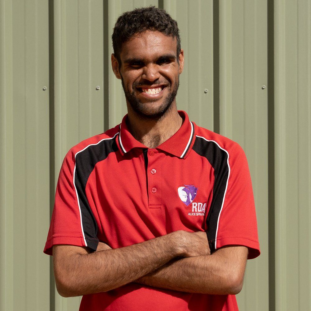 Man with dark hair and a beard wearing a red polo shirt with arms crossed, smiling in front of a green wall.