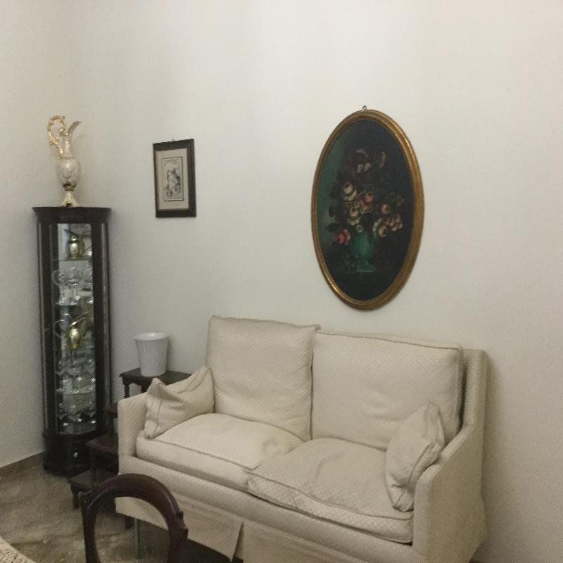 Divano beige in un soggiorno con quadri alle pareti, una vetrina e una piccola lampada da comodino.
