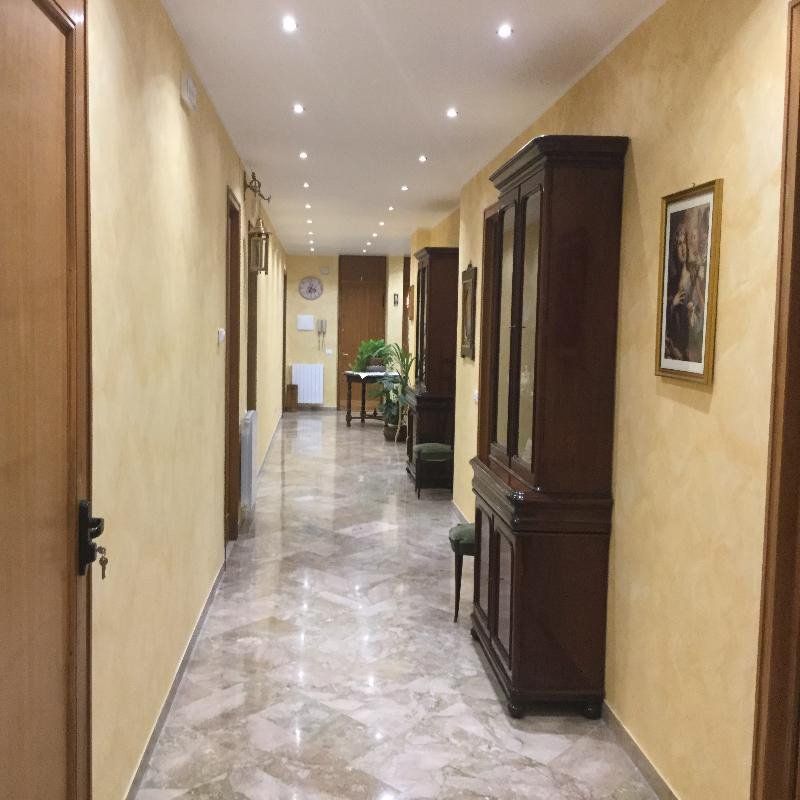Lungo e stretto corridoio con pavimento in marmo, pareti beige, faretti a incasso, armadio e sedia in legno.