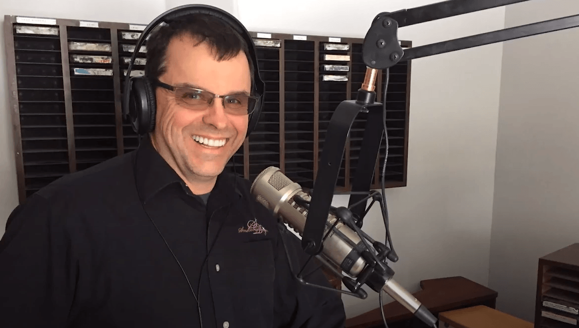 Dr. wearing headphones and glasses is standing in front of a microphone.