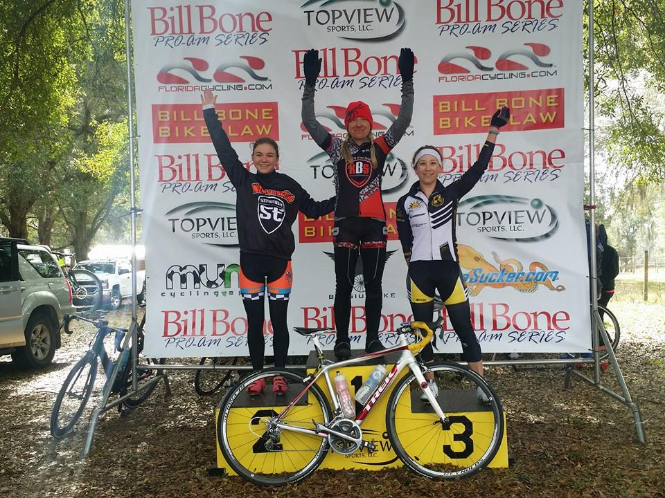 Three women standing on a podium with bicycles in front of a bill bone banner