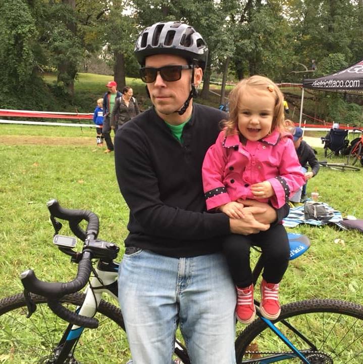 A man wearing a helmet and sunglasses holds a little girl on a bike
