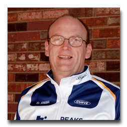 A man wearing glasses and a curve jacket smiles in front of a brick wall