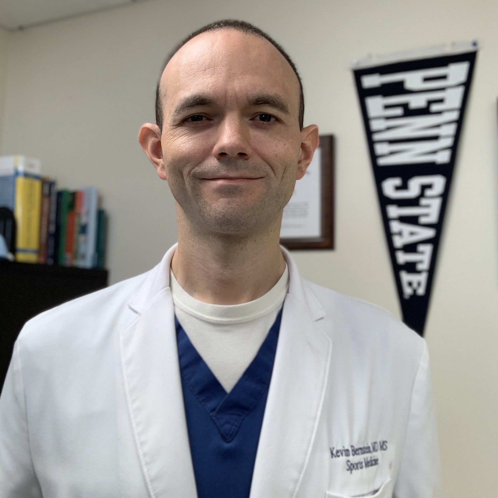 A man in a lab coat is smiling in front of a penn state flag