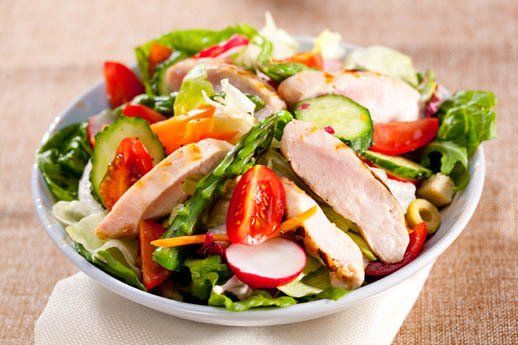 A bowl of salad with chicken , tomatoes , cucumbers , and radishes on a table.