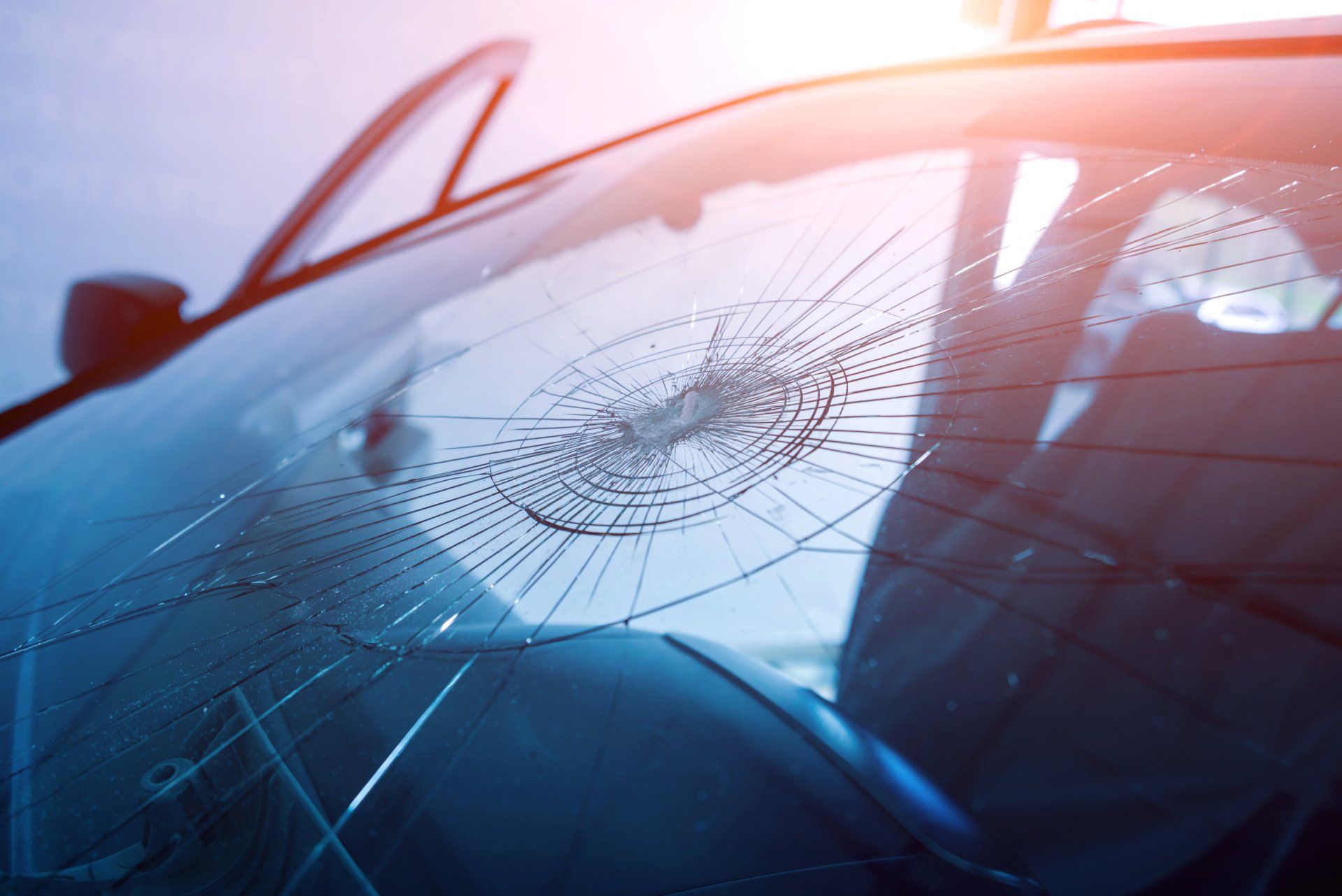 Pare-brise de voiture fissuré avec des fractures en forme de toile d'araignée, reflet du soleil.
