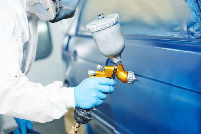 Worker spray-painting a blue car with a paint gun and respirator mask