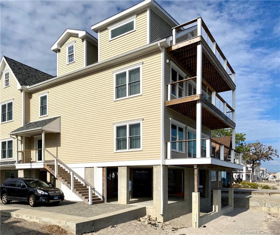 Beige multi-story house with decks, elevated on stilts, steps lead up to entrance, parked car.