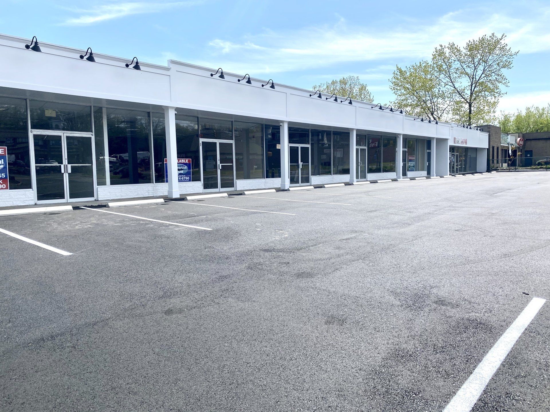A row of commercial buildings with glass storefronts and an empty parking lot on a sunny day.
