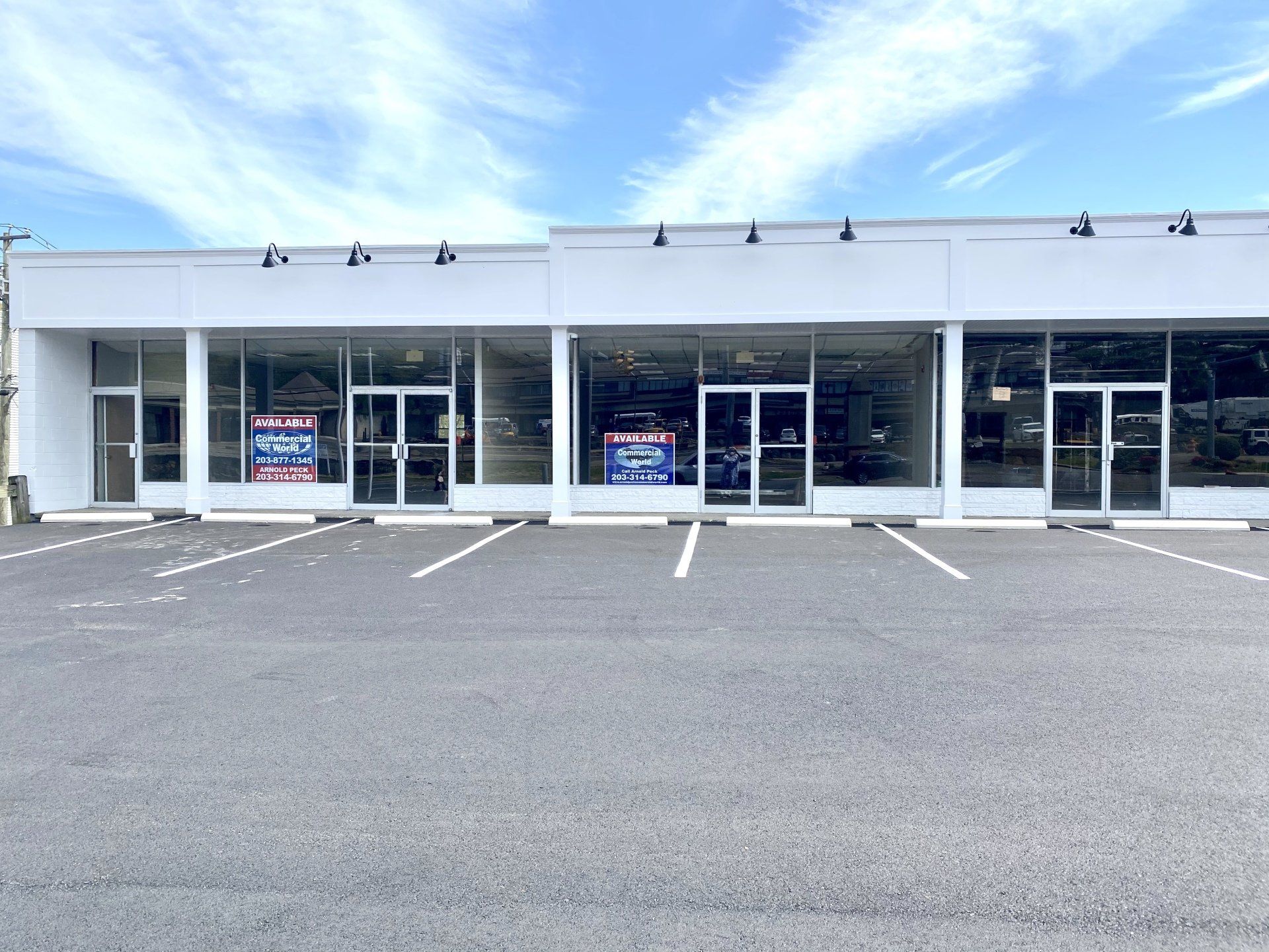 Row of empty storefronts with large windows, under a blue sky, and a paved parking lot.