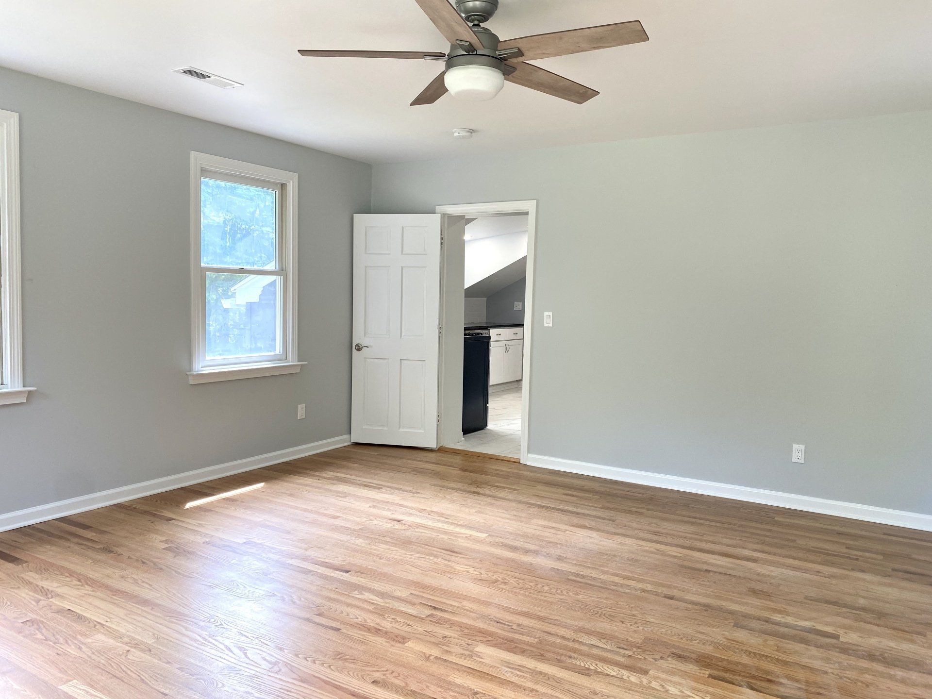 Empty room with hardwood floors, gray walls, open doorway to kitchen, ceiling fan.