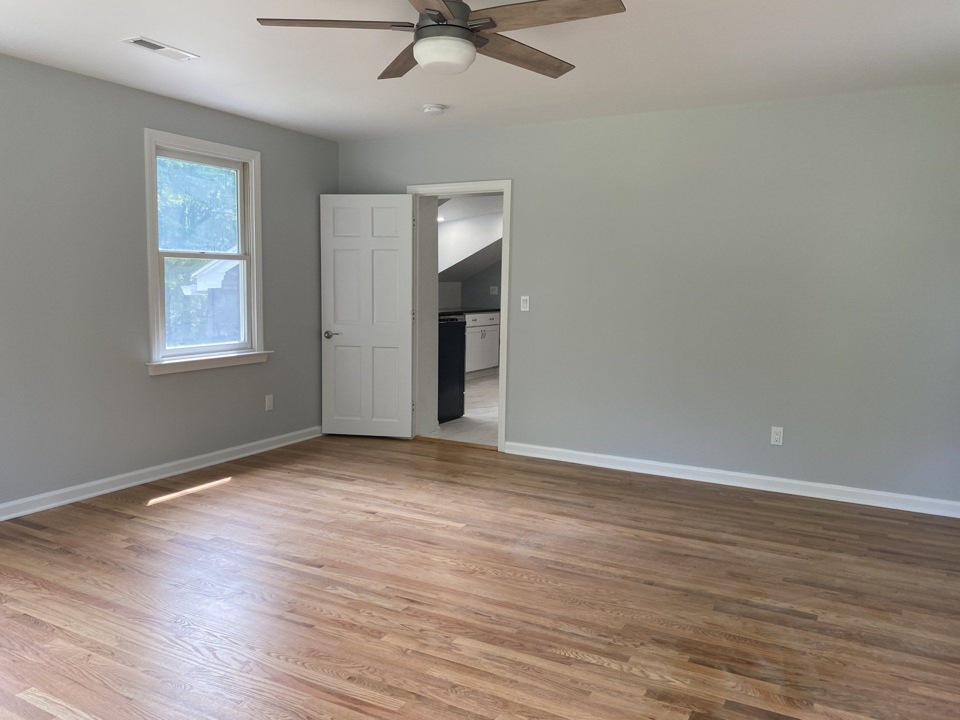Empty room with hardwood floors, a closed white door, and a doorway leading to a kitchen. Light gray walls.