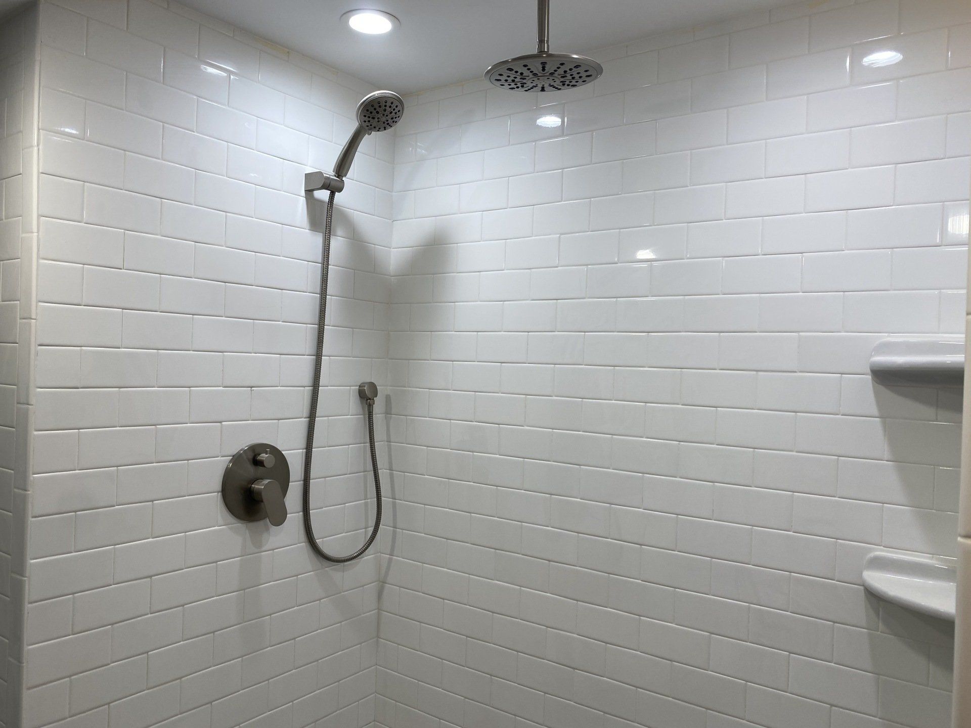 White tiled shower with a handheld and overhead shower head, in a bathroom.