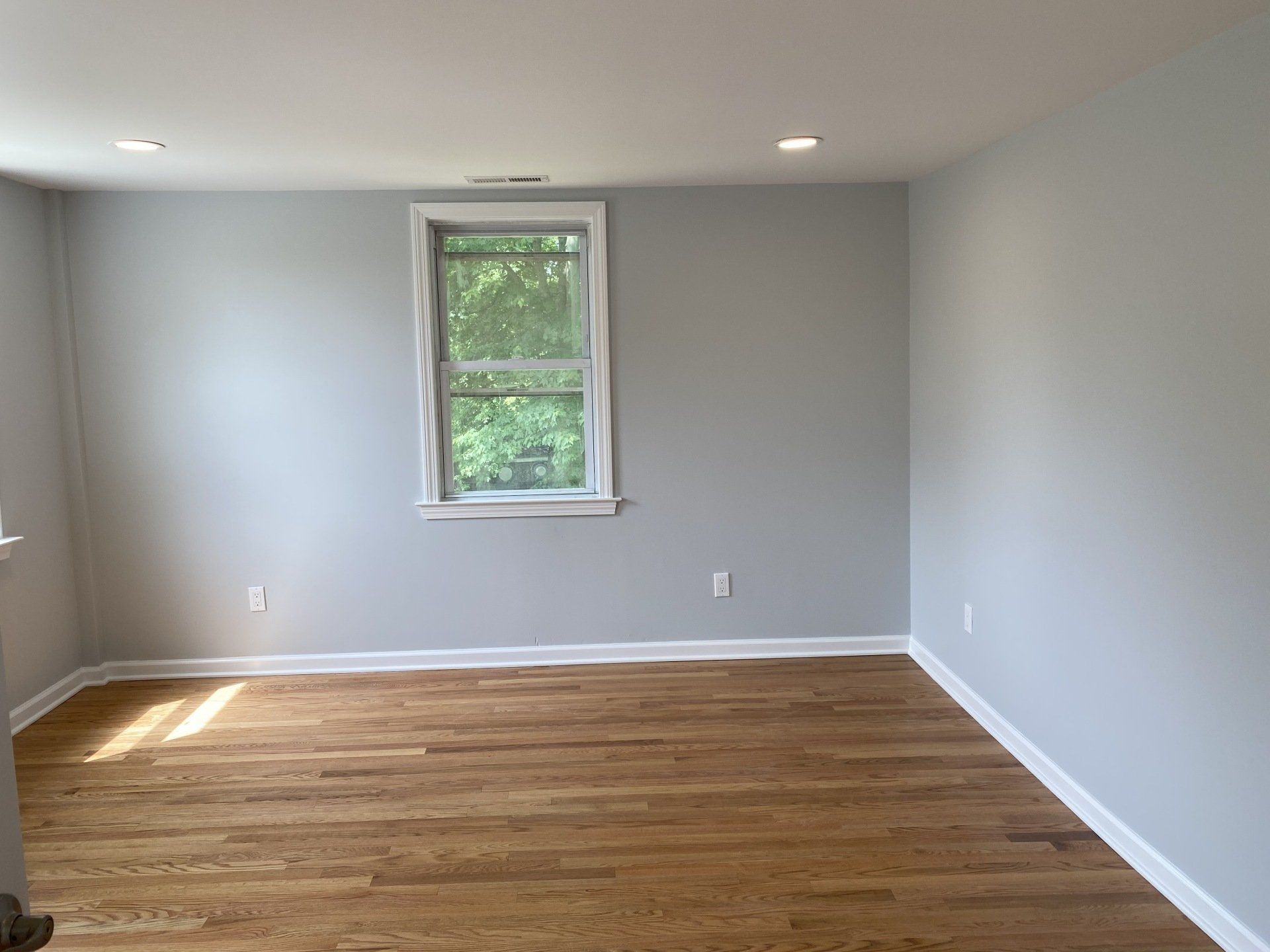 Empty room with light blue walls, hardwood floor, and window.