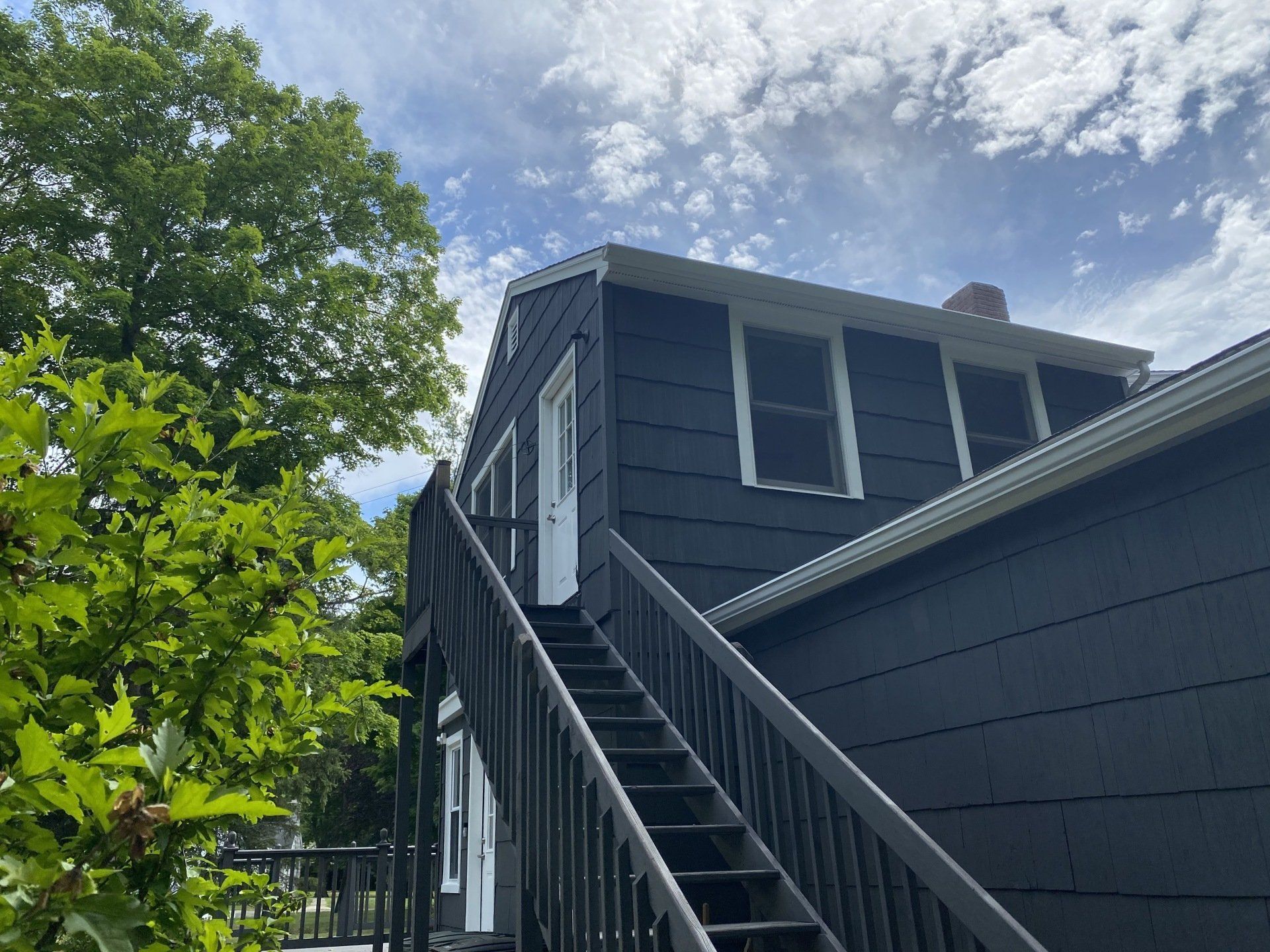 Exterior view of a dark blue house with a staircase leading to a second-floor door, partly obscured by trees.