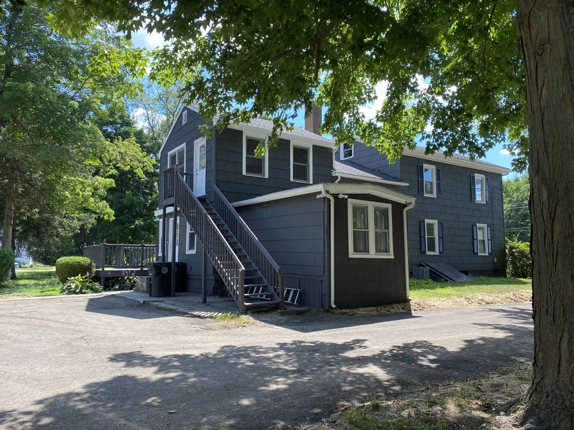 Two-story dark-painted house with exterior stairs, a gravel driveway, and a tree in front.