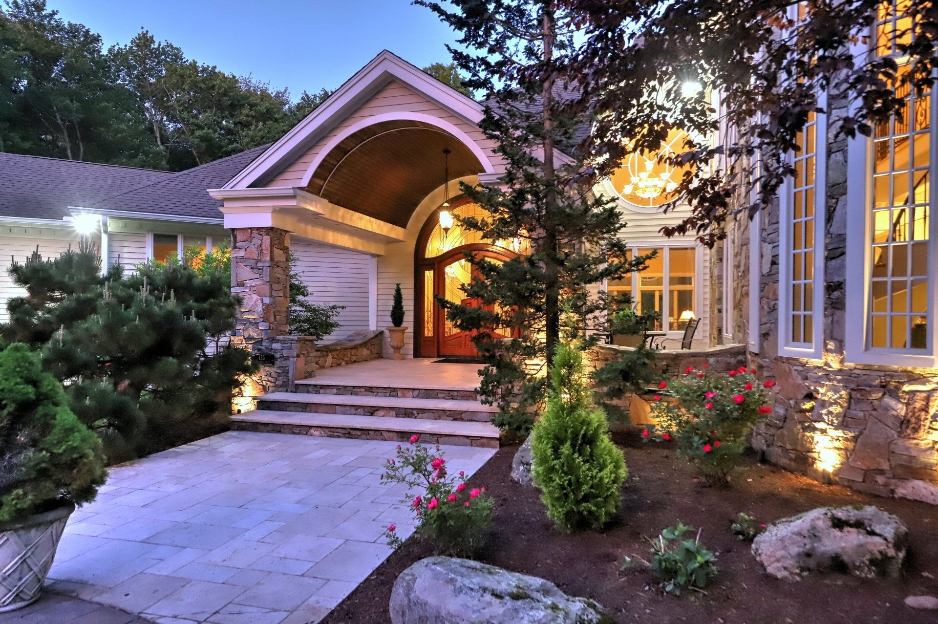 Luxurious home exterior with stone accents, arched entryway, and landscaped garden illuminated at dusk.