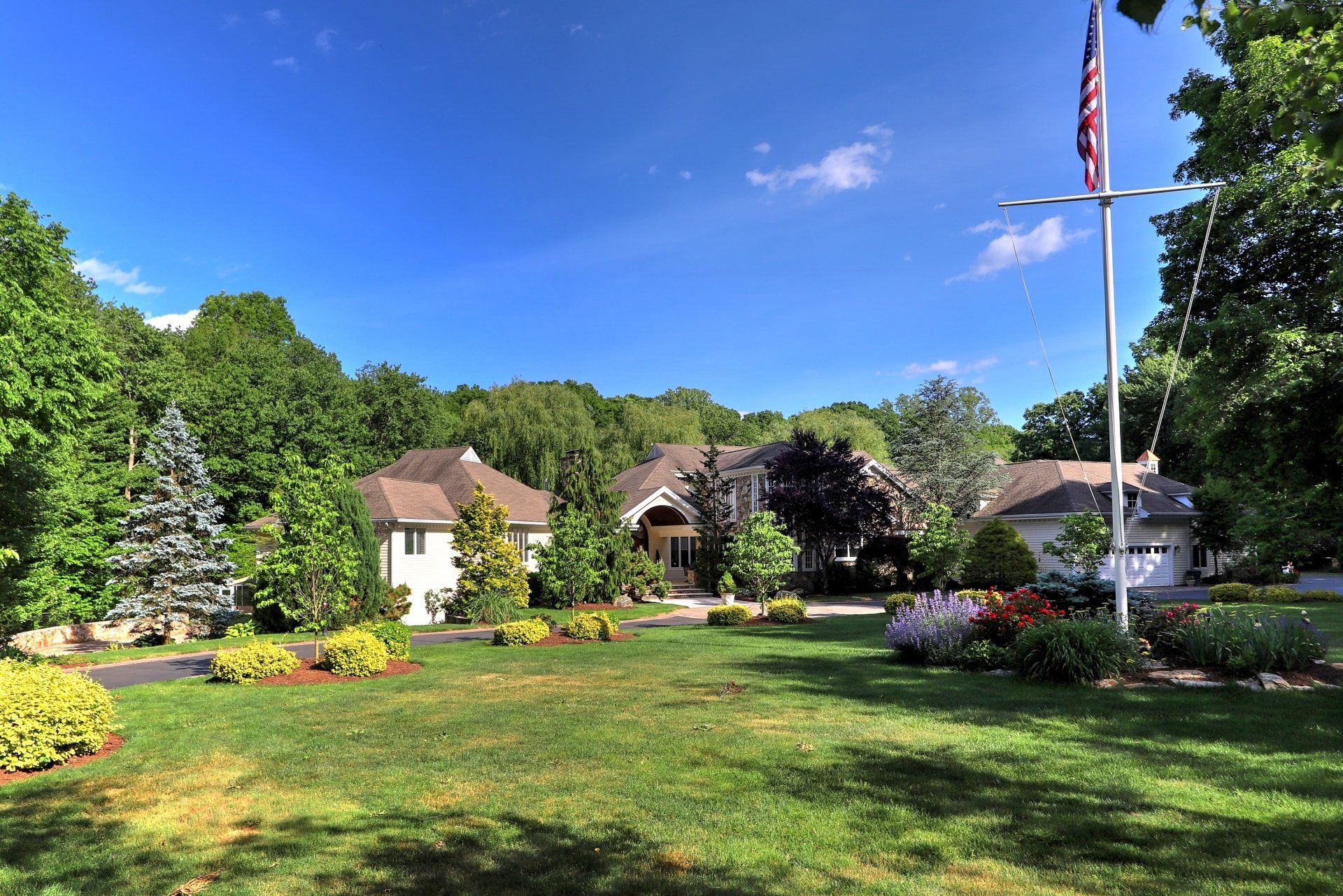 Large house with lush green lawn and flowerbeds, tall flagpole with American flag.
