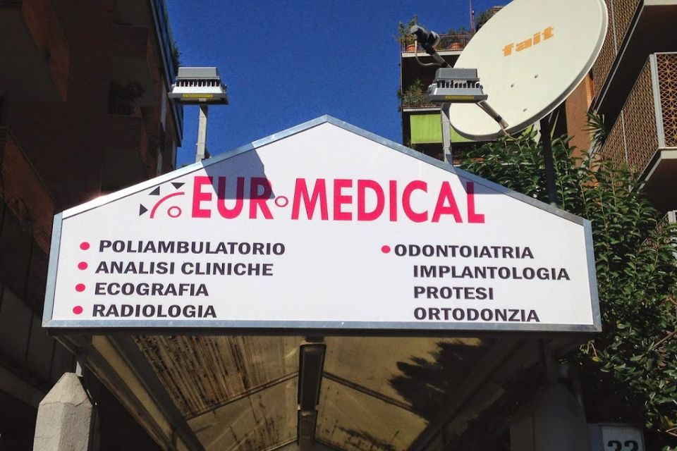 Cartello per il centro medico EUR-MEDICAL. Bianco con testo rosso, elenca servizi come radiologia e odontoiatria. Luce solare, edifici visibili.
