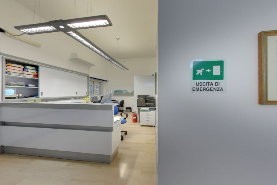 Ufficio con scrivania bianca, segnaletica di uscita di emergenza e illuminazione fluorescente.