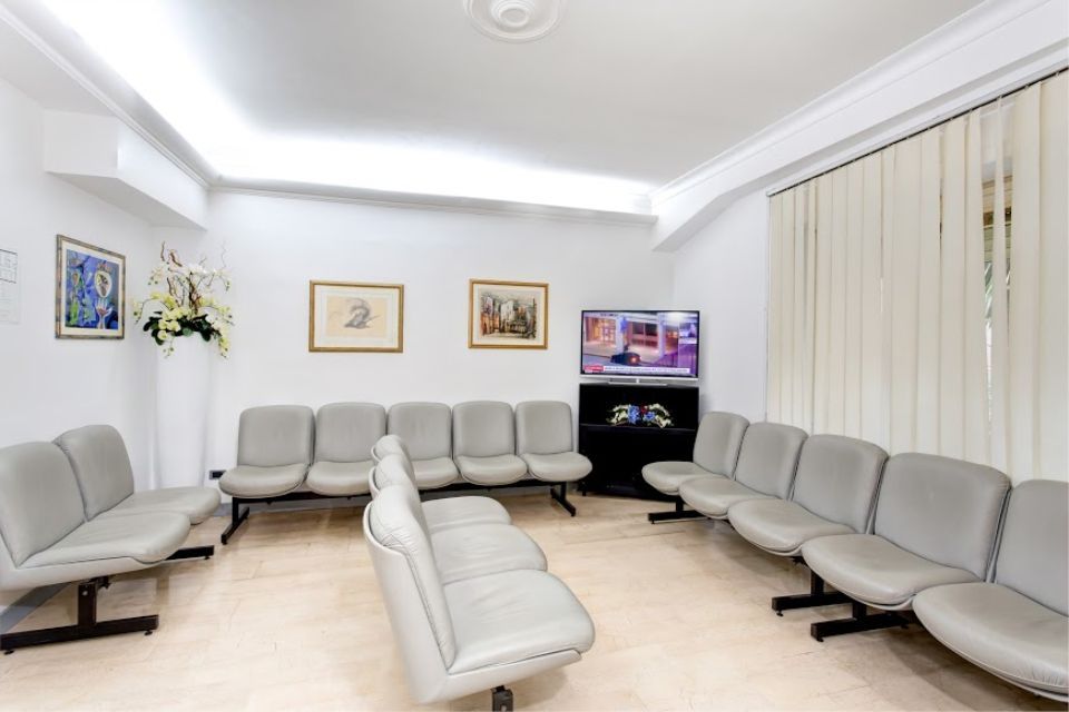 Una sala d'attesa con sedute grigie, una TV e quadri incorniciati su una parete bianca.
