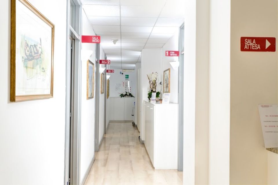 Corridoio lungo e luminoso in una clinica con quadri incorniciati, segnaletica direzionale e pavimenti in legno chiaro.