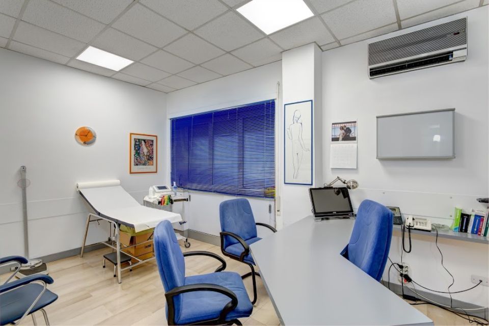 Sala visite del medico con tavolo operatorio, sedie blu, scrivania e tapparelle.