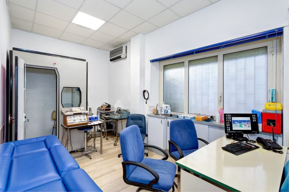 Sala visite mediche con sedie blu, apparecchiature diagnostiche e un lettino per la visita del paziente.
