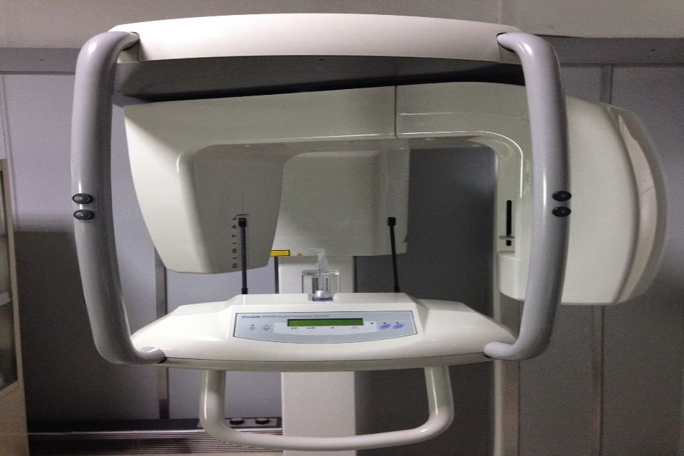 Apparecchio radiologico panoramico dentale; bianco, con bracci curvi e supporto per la testa del paziente.