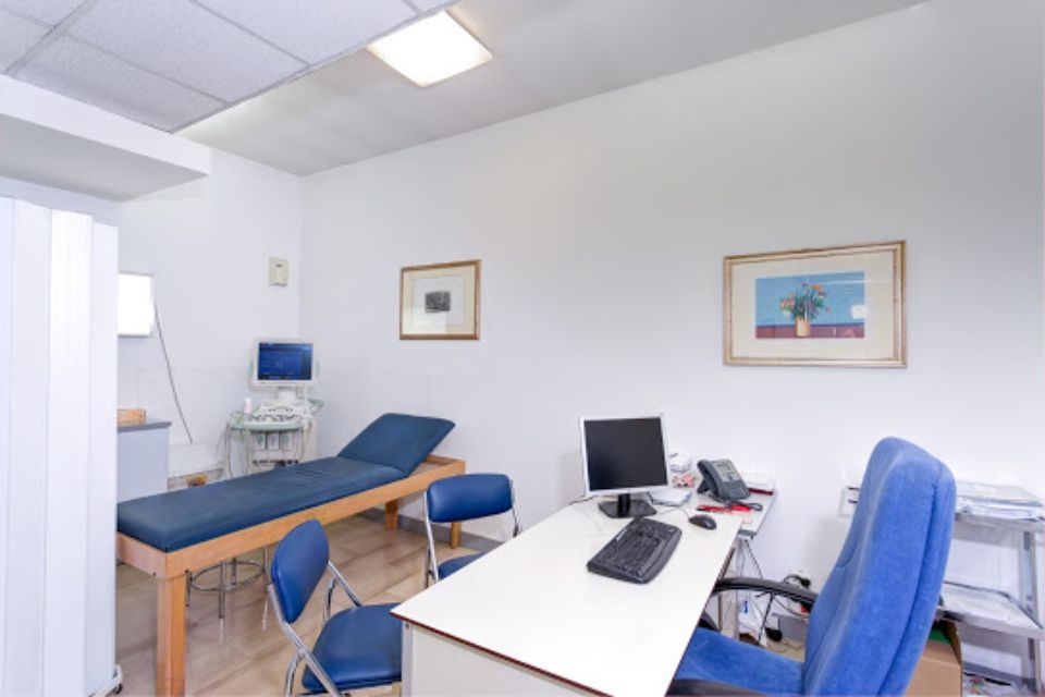 Studio medico: lettino per visite, scrivania con computer, ecografo, sedie blu, quadri incorniciati su pareti bianche.