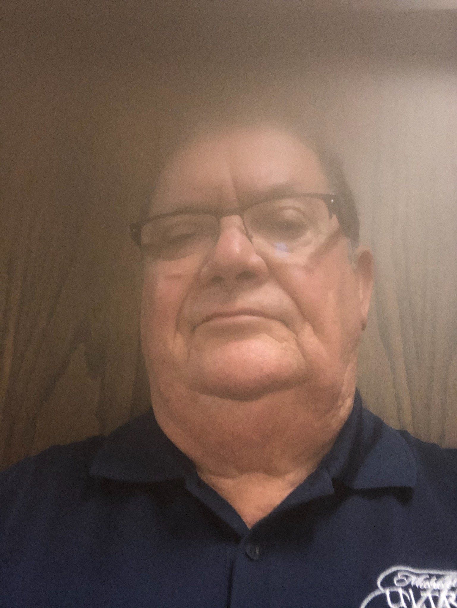 Man with glasses, wearing a dark blue polo shirt, looking at the camera with a neutral expression.
