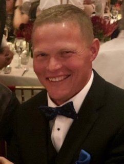 Smiling man in a black tuxedo, blue bow tie, seated at a formal table.