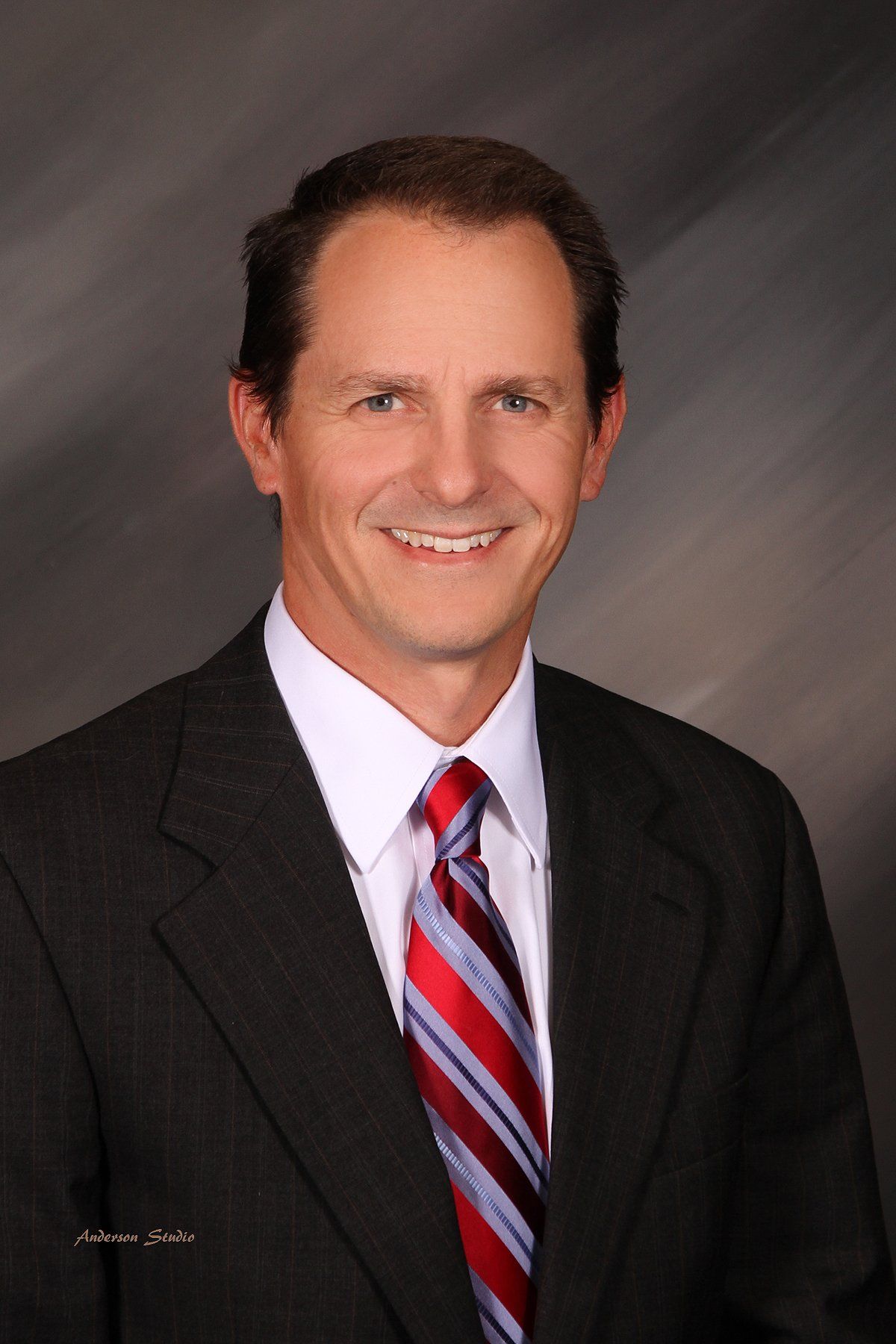 Man in suit smiling, red and blue striped tie, against a gray gradient backdrop.