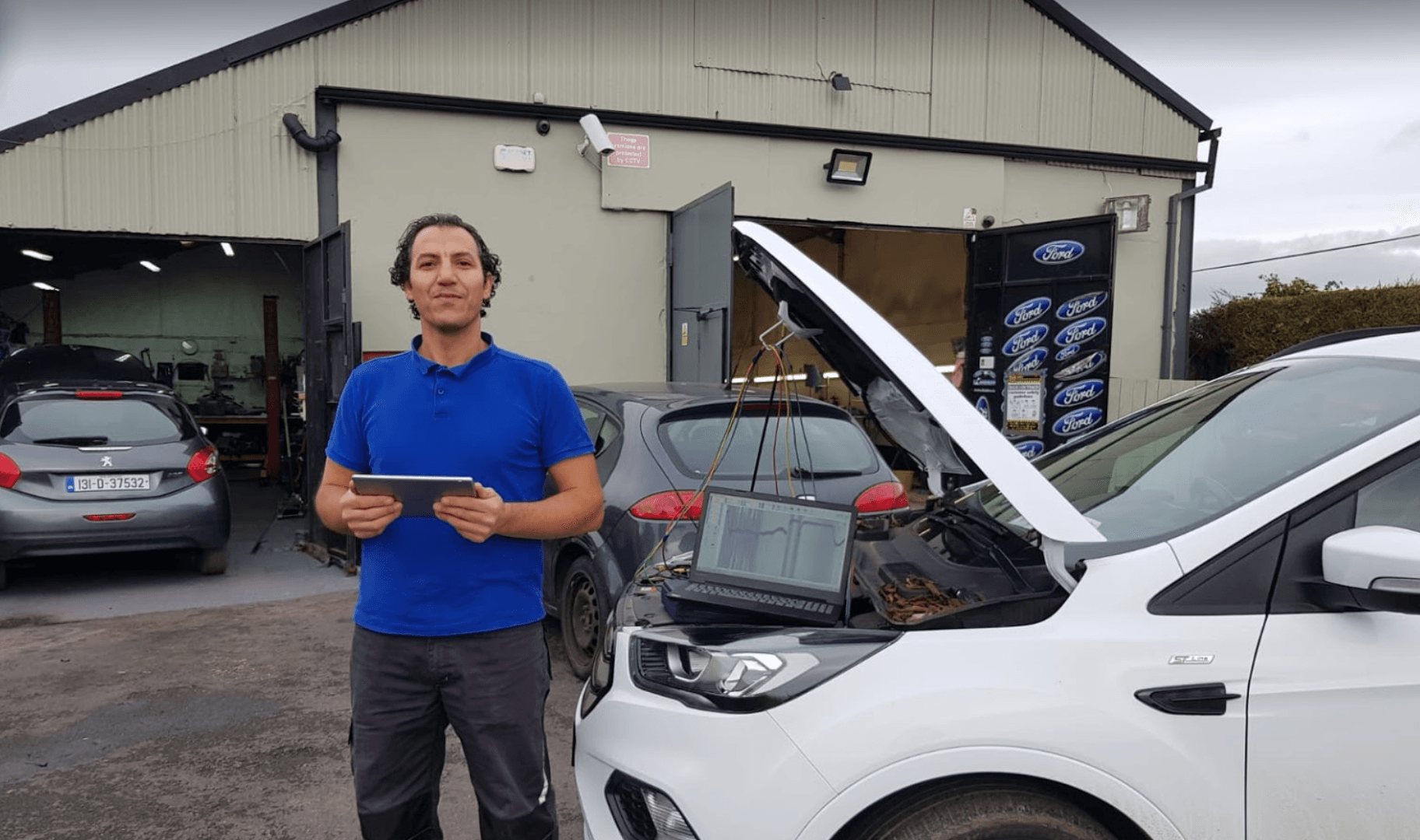 A person in a blue shirt holds a tablet outside a workshop next to a white car with its hood open and a laptop inside.