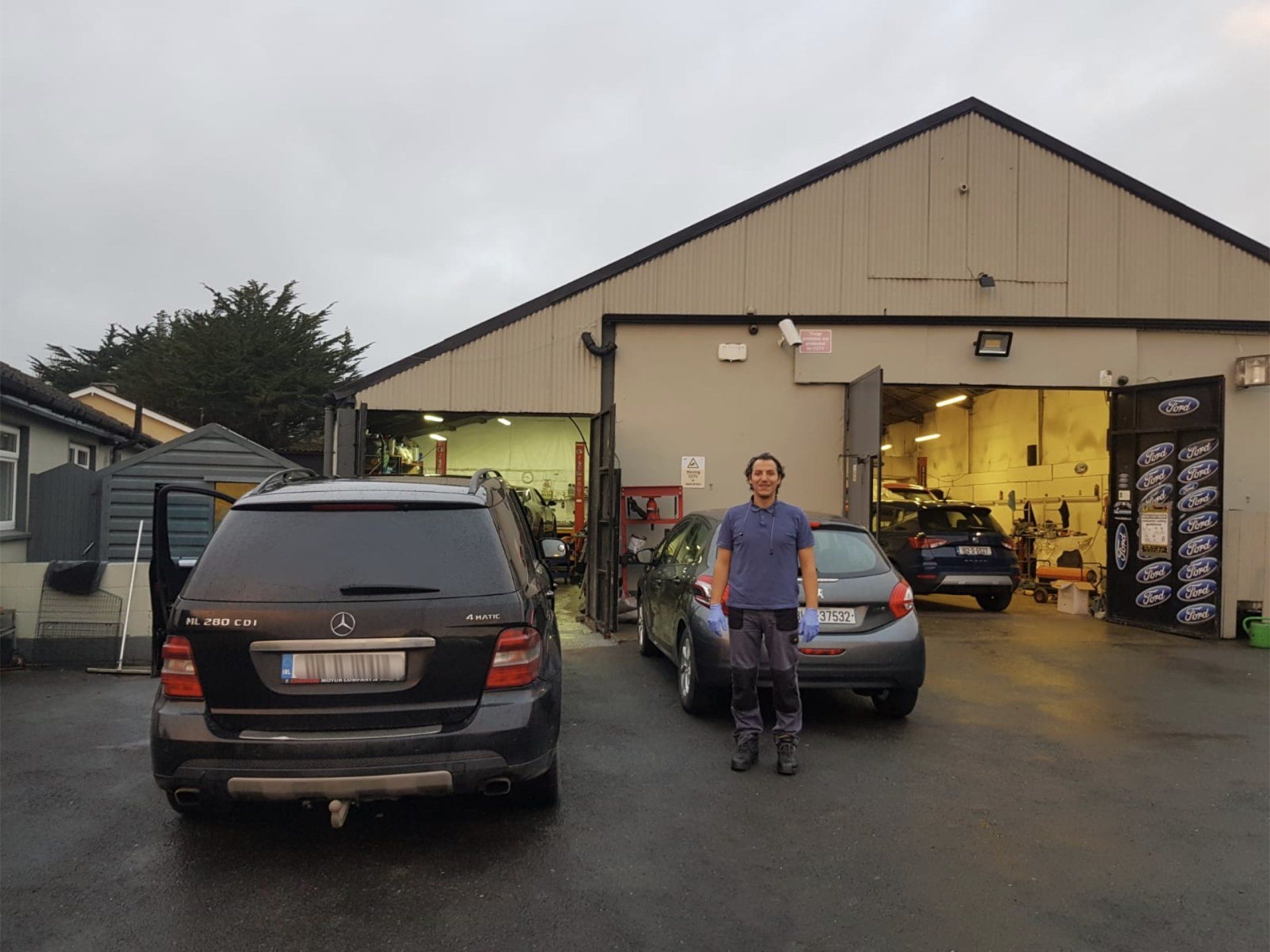 A mechanic in workwear stands outside a workshop garage between a parked black SUV and a grey hatchback.