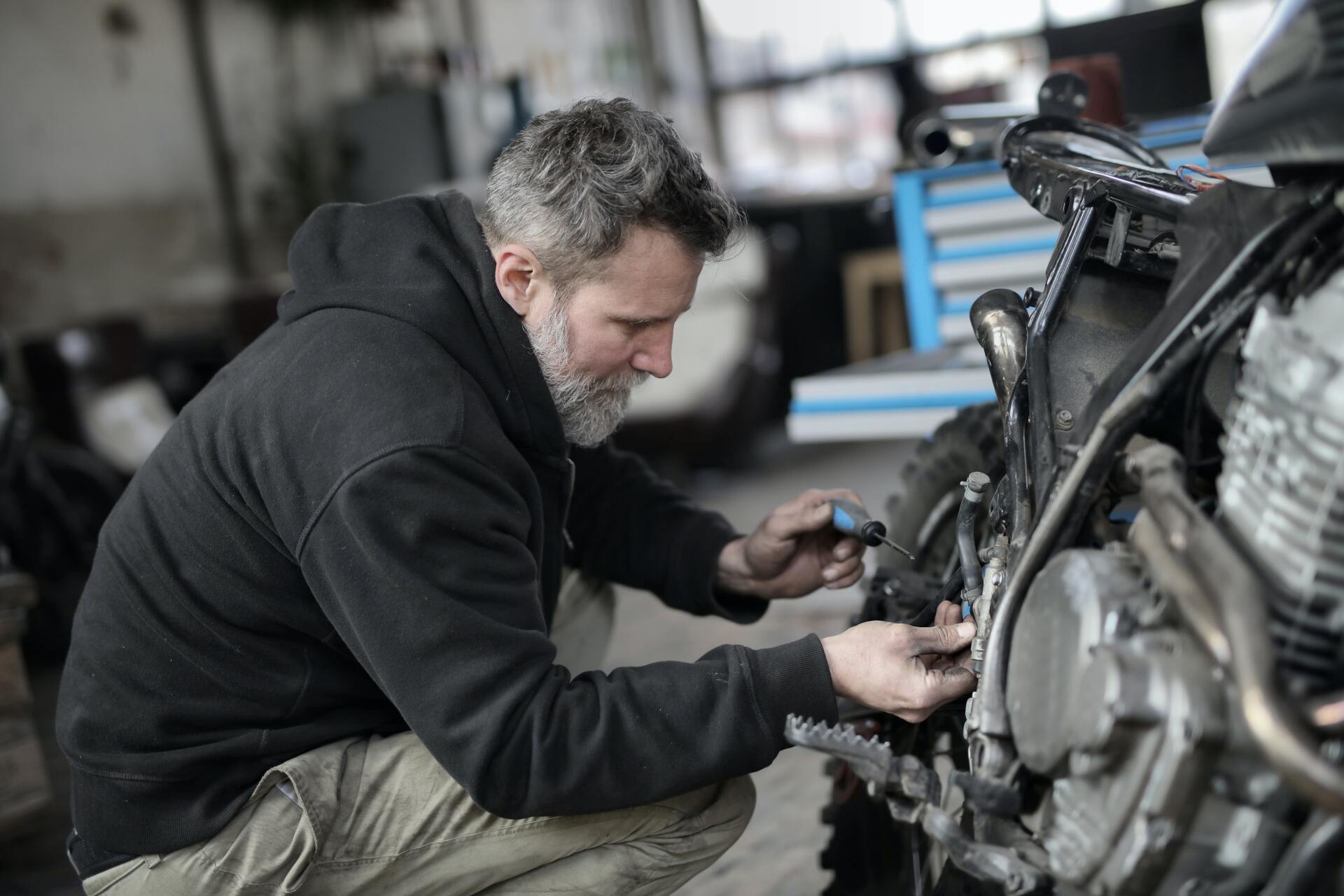 A focused person in a dark hoodie kneeling and inspecting the engine of a motorcycle in a workshop.