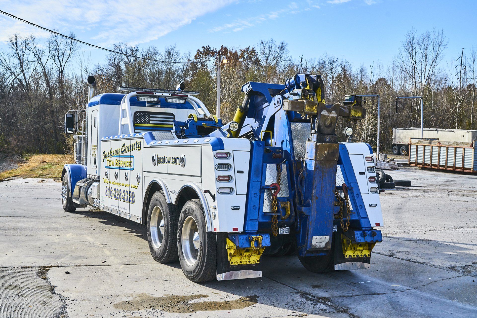 Blue tow truck parked in a lot, lifting equipment extended, with leafless trees in the background.