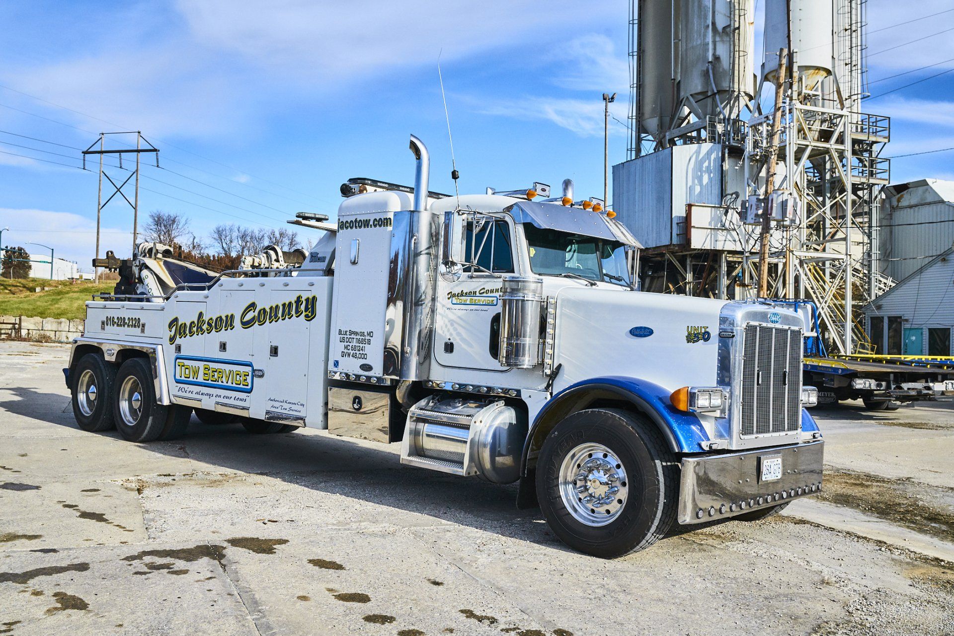 White tow truck parked in an industrial yard beside metal silos on a sunny day