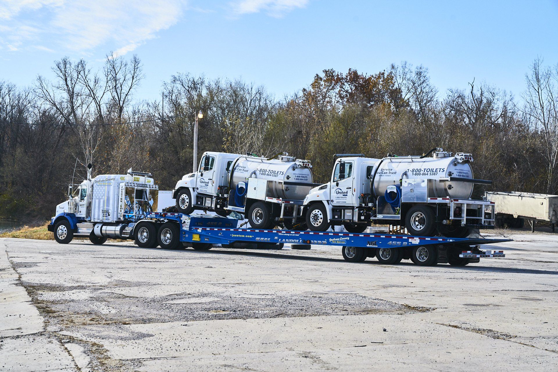 Three white utility trucks loaded on blue trailers in a gravel lot under a clear sky