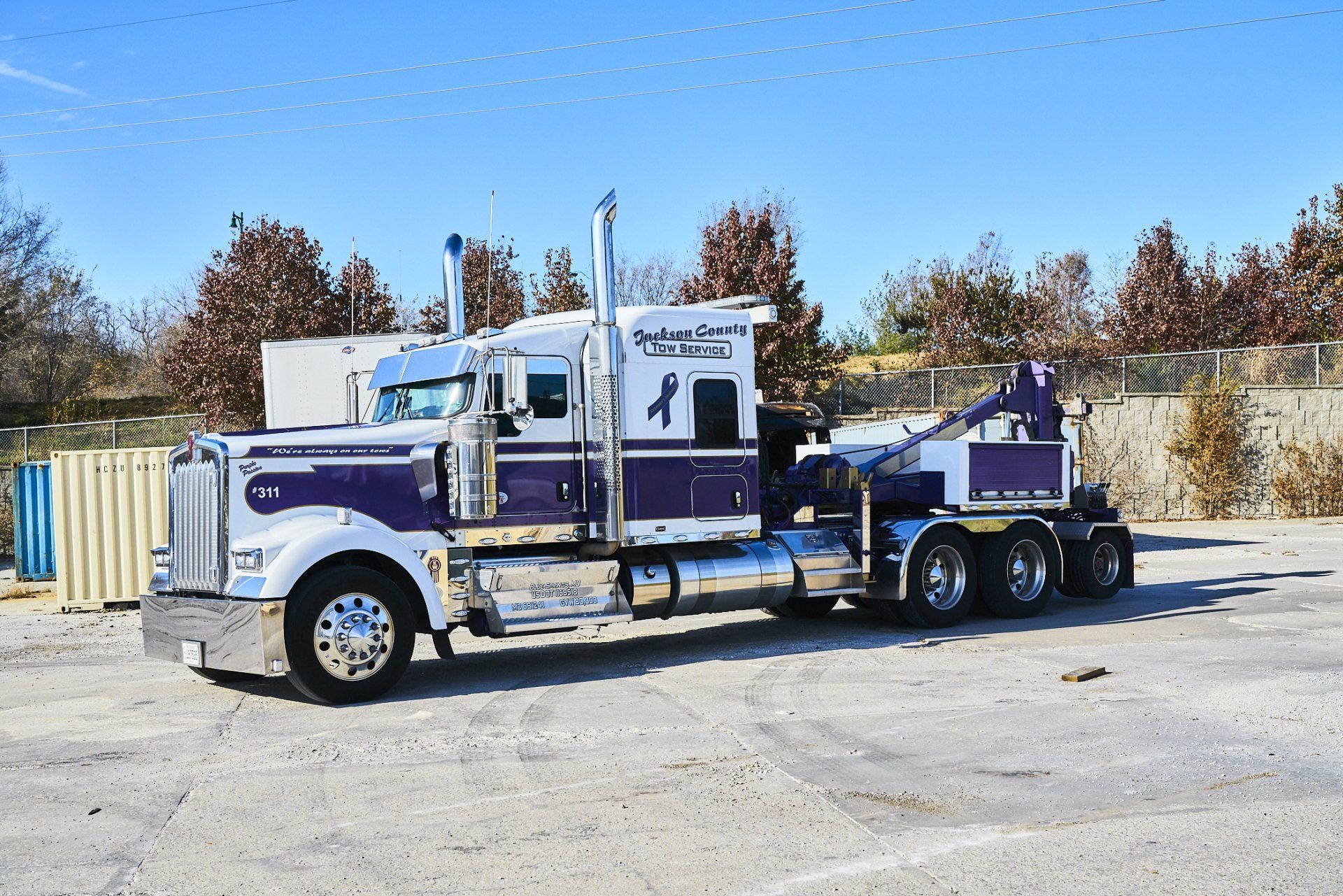 Purple and white semi-truck with a trailer parked on a gravel lot under a blue sky.